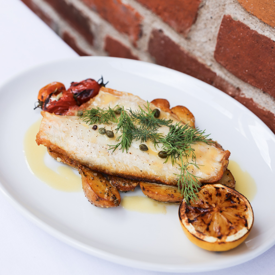 Mediterranean pan-seared fish with roasted tomatoes, potatoes, dill, capers, and a grilled lemon.
